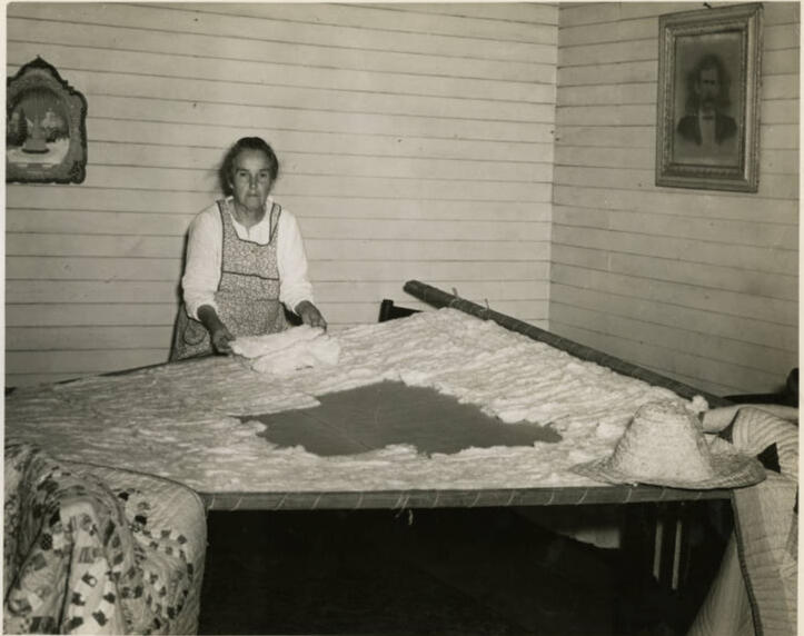 Nancy Page (an old woman in a house with white walls) is stuffing a stretched quilt with cotton.
